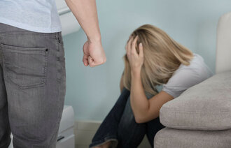 Bildet viser en mann som står med knyttneven foran en redd dame som har krøket seg sammen bak sofaen.