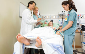 Bildet viser en pasient som ligger i en sykehusseng mens to sykepleiere og en lege står ved siden av. Legen skriver på et skjema.