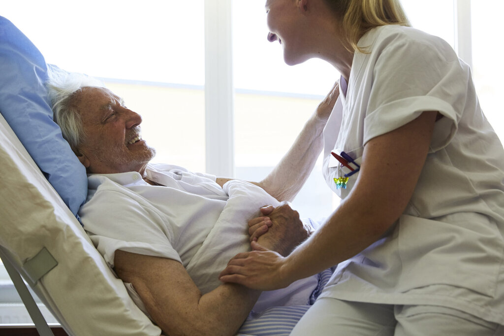 Bildet viser en pasient som ligger i senga, og sykepleieren sitter på sengekanten og holder ham på armen. Begge ler.