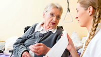 Bildet viser en eldre dame hjemme hos seg selv, og en sykepleier som ruller ut en bandasje mens hun snakker med pasienten. 