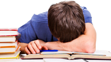 Bildet viser en fortvilet student som hviler hodet på pulten mens han er omgitt av bøker