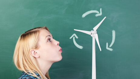 Bildet viser en jente i profil foran en tavle. Hun blåser på en krittegning av en vindturbin
