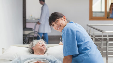 Bildet viser en sykepleier som lener seg over en pasient mens hun smiler til fotografen