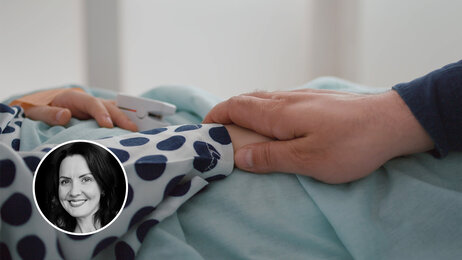 Bildet viser et utsnitt av en voksen hånd som holder rundt en barnehånd. Barnet ligger i sengen.