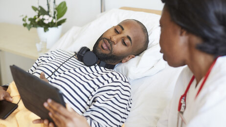 Bildet viser en pasient som ligger i sengen og ser på en håndholdt skjerm, mens en sykepleier sitter på sengekanten