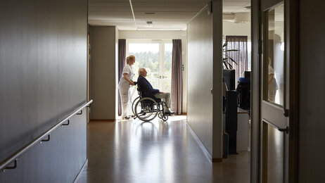 Bildet viser en sykepleier som triller en pasient i rullestol på et bo- og behandlingssenter