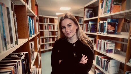 Bildet viser Cathrine Emilie Valgermoe Forren. Hun står i et bibliotek, ser mot kamera og har armene i kors.
