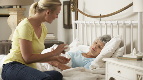Bildet viser en eldre, syk dame i senga. På sengekanten sitter dattera