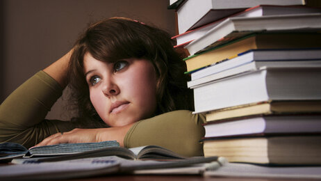 Bildet viser en student med et litt oppgitt uttrykk, som ligger over en oppslått bok på pulten. Det ligger en stabel bøker ved siden av henne.