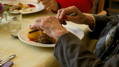 Bildet viser et middagsbord på sykehjem med to middagstallerkner og hender til to gamle personer