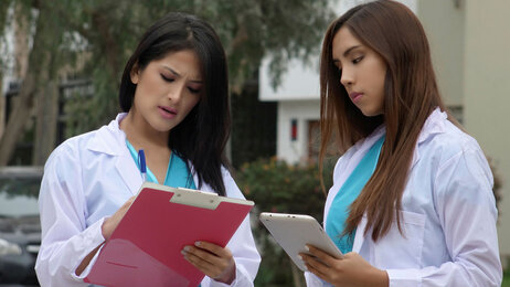 Bildet viser to kvinner med sykepleieruniform som står og snakker sammen utendørs. Den ene holder en mappe, mens den andre noterer på en skriveblokk.
