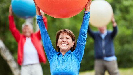 Bildet viser en smilende eldre kvinne i treningstøy som holder en stor ball over hodet.