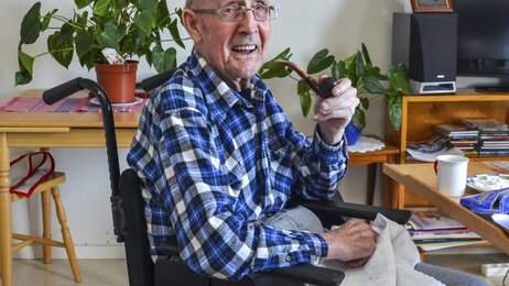 Bildet viser John Gundersen på 93 år. Han sitter i en rullestol i omsorgsboligen sin. Han har en pipe i hånden og smiler til fotografen.