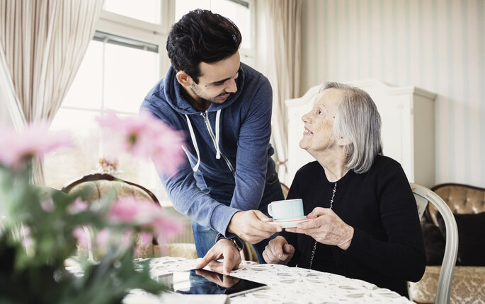 Bildet viser en pleier som gir en smilende, eldre pasient en kopp kaffe