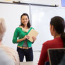 Bildet viser ei dame som står foran en whiteboard og underviser. Hun holder en skriveblokk i hånda. I rommet ser vi to voksne, sittende damer bakfra