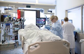 Bildet viser en intensivavdeling, der en pasient ligger i sengen mens en sykepleier står ved siden av. I bakgrunnen står legen og sjekker på en skjerm