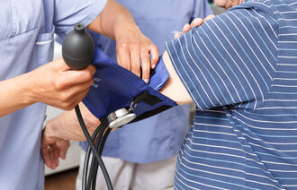 Bildet viser en sykepleier som måler blodtrykket på en eldre mann. En annen sykepleier skimtes i bakgrunnen