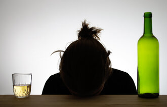 Bildet viser silhuetten av en kvinne som hviler hodet på en bordkant. Ved siden av henne står et glass med alkohol og en flaske.
