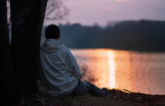 Bildet viser en ung mann sett bakfra som sitter ved et tre og ser ut på en solnedgang over et vann