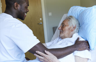Bildet viser en mannlig sykepleier som smiler og holder hånden sin på skulderen til en liggende pasient