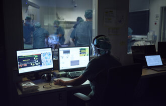 Bildet viser en operatør av en simulering, som sitter og styrer det hele via tre dataskjermer, foran et ensidig speil. På andre siden foregår simuleringen