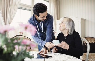 Bildet viser en pleier som gir en smilende, eldre pasient en kopp kaffe