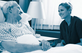 The photo shows a male older patient lying in a hospital bed. A female nurse is sitting beside him. Both are smiling