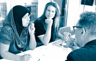 Bildet viser en dame med hijab, en dame med vestlige klær og en mann som sitter rundt et bord. De har PC og skrivesaker foran seg og deltar i møte.