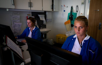 Bildet viser to sykepleiere som sitter foran hver sin datamaskin