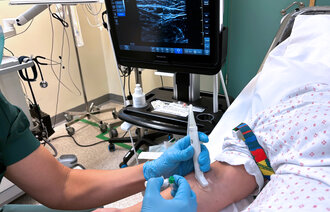 The photo shows a nurse using ultrasound to locate a vein before catheterisation