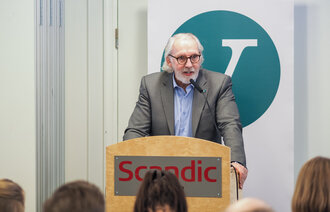 Senior mannlig politiker på talerstol i stort lokale med Venstres logo i bakgrunnen