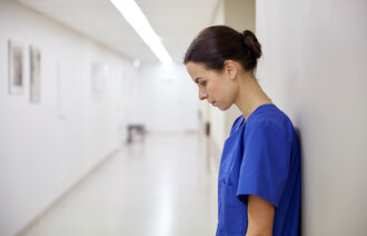 Kvinnelig helsearbeider står inntil veggen i en sykehuskorridor og kikker i golvet. Hun virker sliten eller fortvilet.