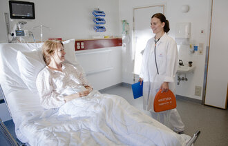 Bildet viser en lege på legevisitt hos en pasient. I hånden holder legen en visittstol