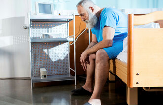 Bildet viser en eldre mann med skjegg som sitter på kanten av en sykehusseng med hendene i fanget.