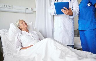 Bildet viser en syk, eldre dame i en sykeseng. Ved sengen står en lege og sykepleier på visitt.