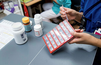 Bildet viser en apoteker/hjemmesykepleier/sykepleier som legger medisiner oppi en dosett. Ved siden av står flere pilleesker.
