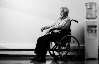 Bildet viser en eldre mann som sitter i en rullestol ved siden av en vanndispenser