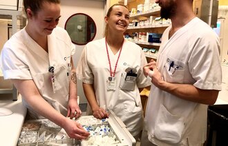 To kvinnlige og en mannlig sykepleier i hvite uniformer i medisinrom. Smiler og ser på små gjenstander i rommet.  
