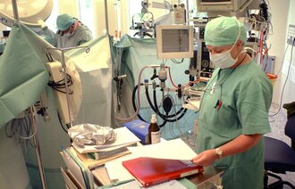 Bildet viser en anestesisykepleier som kikker i en perm. Bak henne ses en kirurg som holder på å operere.