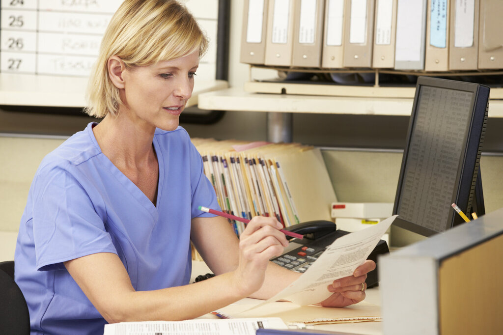 Bildet viser en sykepleier som sitter ved en pult og gjør papirarbeid