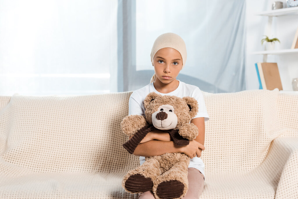 Bildet viser et sykt barn som sitter i en sofa og har en teddybjørn i fanget