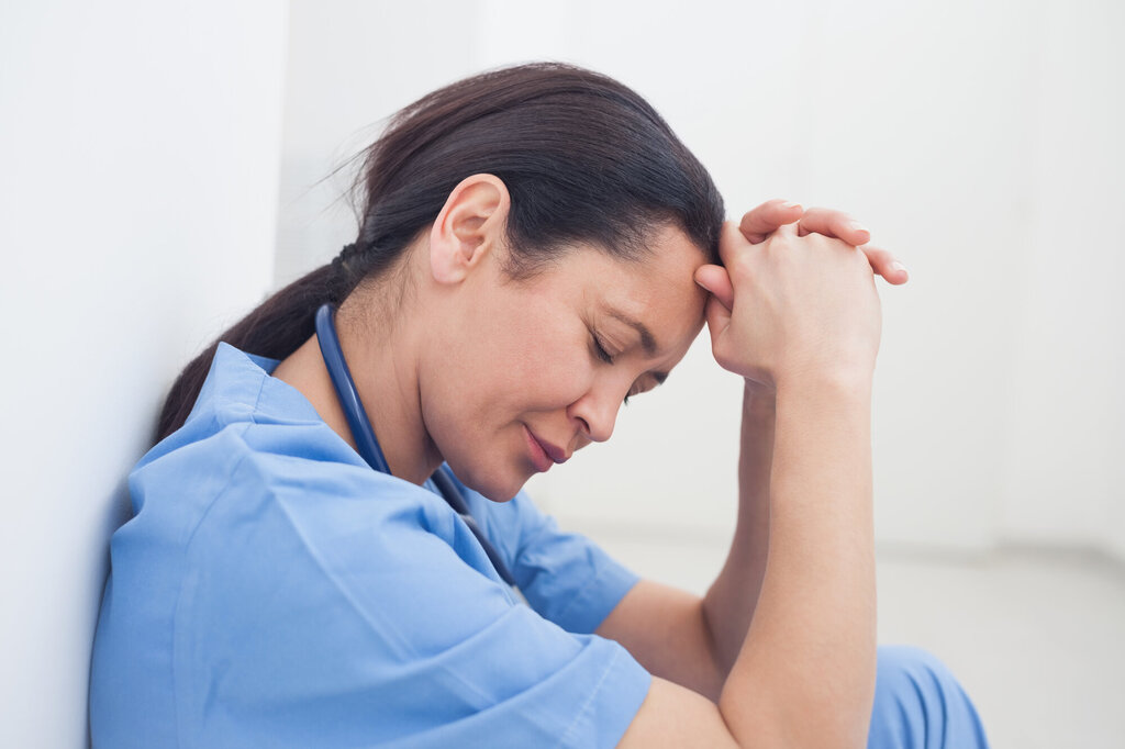 Bildet viser en fortvilet sykepleier som sitter i ganga og kikker nedover, mens hun lener hodet mot foldede hender