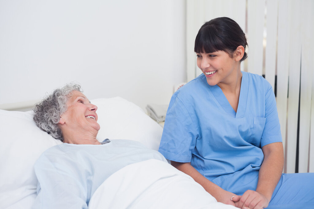 Bildet viser en eldre kvinne som ligger i en seng og smiler til et yngre kvinnelig helsepersonell som smiler tilbake