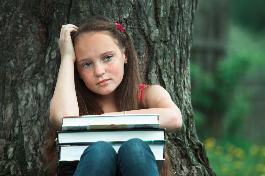 Bildet viser en ung jente som sitter ved et tre med en stabel bøker på knærne