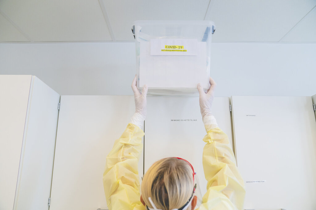 Bildet viser en sykepleier i gul smittefrakk som løfter en boks ned fra toppen av et skap.