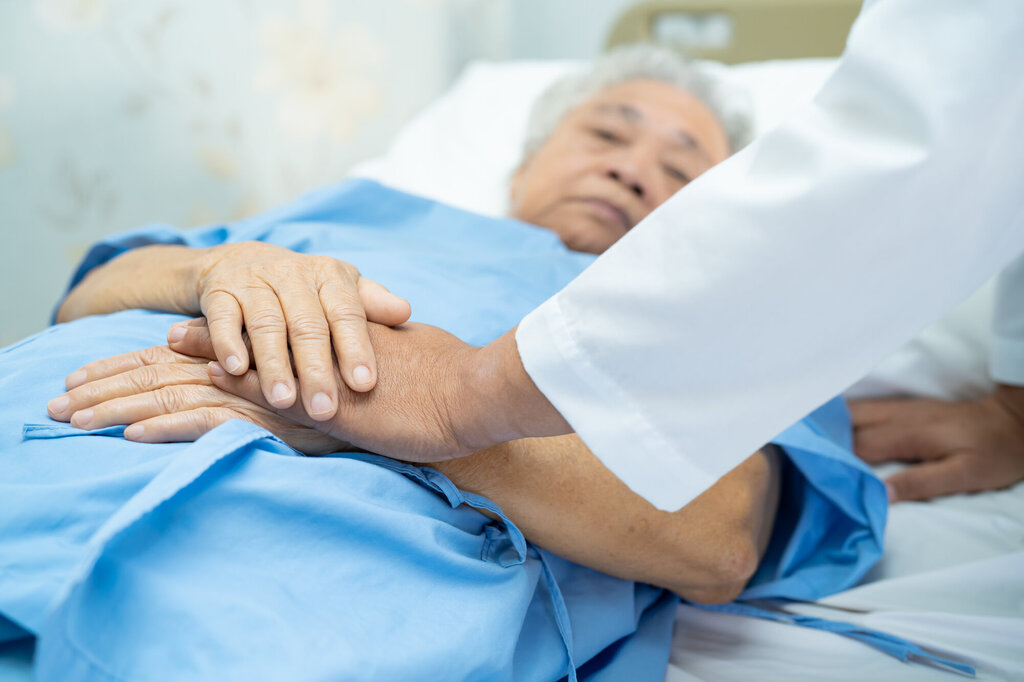 Bildet viser en eldre mann som ligger i en seng, og et helsepersonell som holder vedkommende i hendene