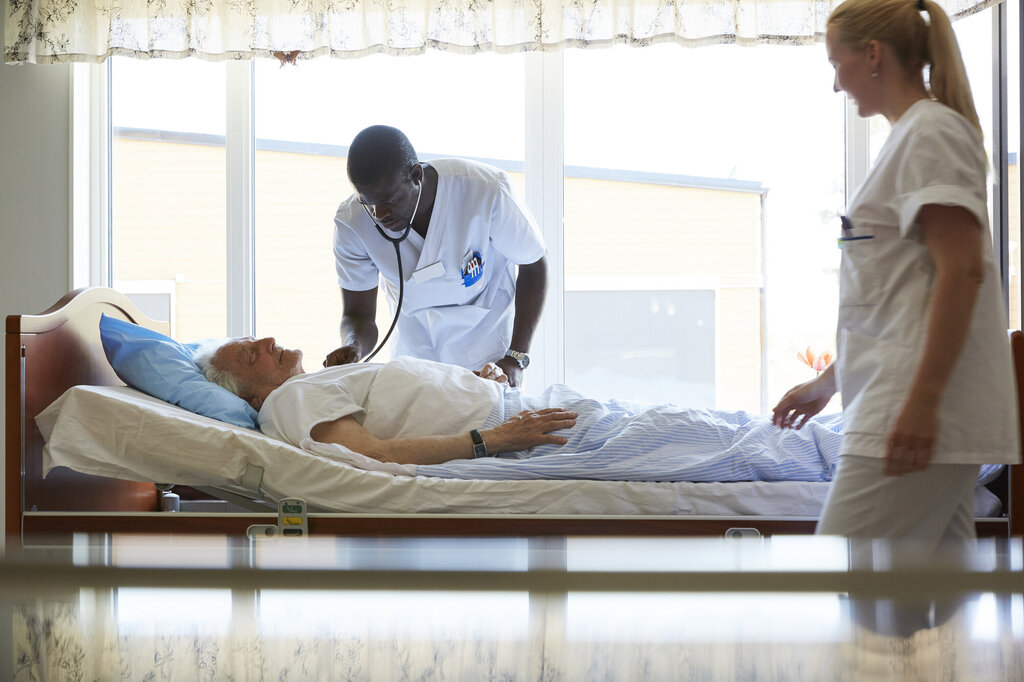 Bildet viser en sykepleier som lytter med stetoskop på en eldre mann i sengen. Ved siden av står en annen sykepleier og observerer.