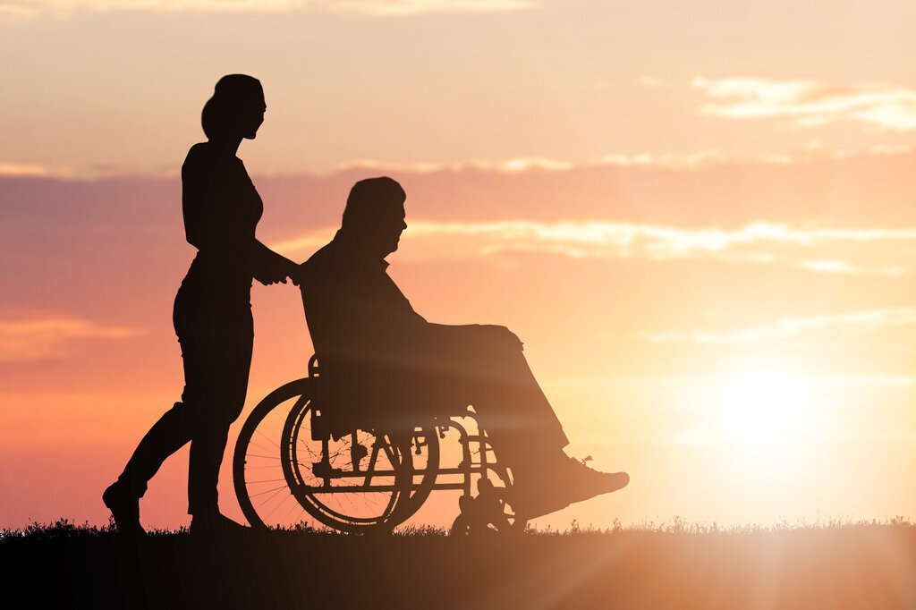 Bildet viser silhuetten av en kvinne som triller en eldre mann i rullestol i solnedgang.