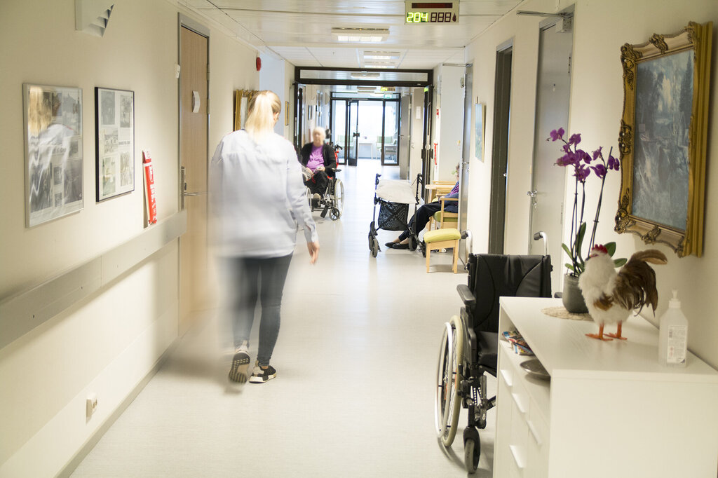 Bildet viser en korridor hvor en sykepleier går mot en eldre pleiepasient i rullestol.