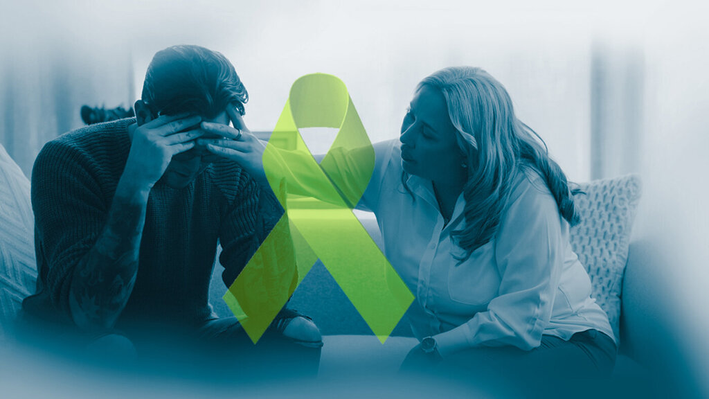 The photo shows a despaired man being comforted by a woman. Between them there is a green ribbon, the symbol of mental health